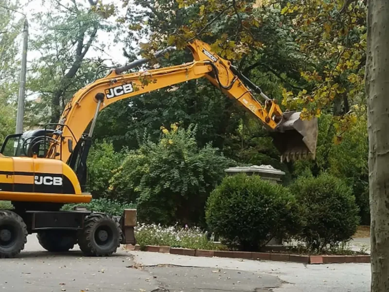 Желтый экскаватор JCB заносит ковш над каменным постаментом демонтированного бюста Мичурина на территории Селекционного института в Одессе